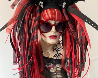 May include: A mannequin head wearing red and black cat-eye sunglasses and a headpiece featuring black feathers, silver skull accents, and long, dangling red and black fabric strips, mesh ribbons, and black leather-like straps with buckle holes.