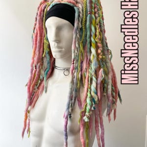 May include: A mannequin head and torso displaying vibrant, handmade dreadlocks. The dreadlocks are a mix of pink, yellow, green, and blue, with a black headband. The text "MissNeedles Hair" is displayed vertically on the right side.