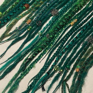 May include: A selection of green and teal dreadlocks, decorated with beads and charms. The dreads vary in shade, with some featuring black stitching and accents. The accessories include silver charms and wooden beads, creating a unique, handcrafted appearance.