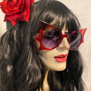 Puede incluir: Una pinza para el pelo de rosa roja con un par de gafas de sol de ojo de gato rojas con montura negra. Las gafas de sol tienen una lente degradada que es más oscura en la parte superior y más clara en la parte inferior.