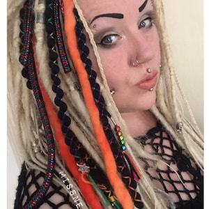 Op de afbeelding: Een persoon met blonde dreadlocks met oranje, rode, zwarte en blauwe accenten. De dreadlocks zijn versierd met kralen en bedels. De persoon draagt een zwart fishnet topje en heeft meerdere piercings in het gezicht.