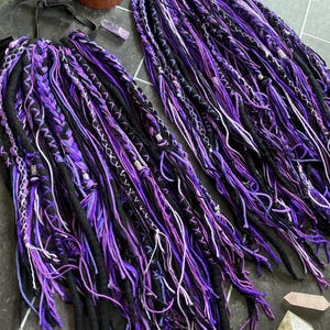 Könnte beinhalten: Zwei Sets handgefertigter Dreadlocks in Schwarz, Lila und Weiß. Die Dreads sind aufwendig mit gewickelten Abschnitten und dekorativen Elementen gestaltet. Die Dreadlocks sind auf einer dunklen Oberfläche angeordnet und zeigen die lebendigen Farbkombinationen.