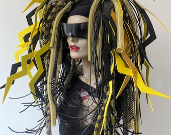 Könnte beinhalten: Ein Schaufensterpuppenkopf mit schwarzer Visier-Sonnenbrille und einem voluminösen Kopfschmuck aus langen, herabhängenden schwarzen und gelben Blitzformen, metallisch goldenen Netzschläuchen und dünnen schwarzen Kordeln in einem dichten, mehrschichtigen Stil.