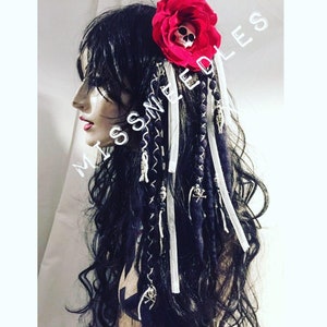 Op de afbeelding: Zwarte en witte dreadlocks met witte lintaccenten en schedelcharms. Een rode roos met een schedel in het midden is bevestigd aan de dreadlocks.