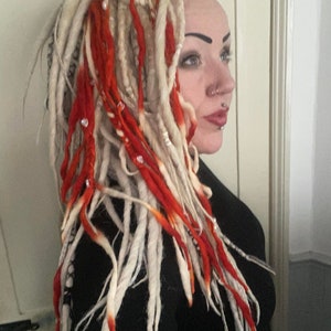 Op de afbeelding: Een persoon met lange, witte dreadlocks met rode accenten. De dreadlocks zijn versierd met kralen en worden in de nek gedragen. De persoon draagt een zwart shirt.