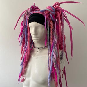 May include: A headpiece featuring long, thick yarn dreadlocks in shades of bright pink, soft lavender, and light blue. The dreadlocks are attached to a black headband worn on a mannequin head, which also features a silver-toned metal collar necklace.