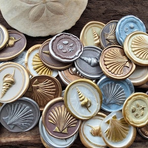 May include: A collection of gold, silver, and white wax seals with various seashell designs. The seals are arranged on a wooden surface.