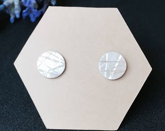 May include: A pair of silver stud earrings with a textured, geometric design. The earrings are displayed on a hexagonal card.