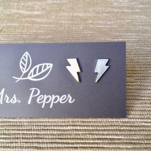 Peut inclure: Une paire de boucles d'oreilles clous en forme d'éclair argentées sur une carte grise avec le texte "Mrs. Pepper" imprimé en blanc.