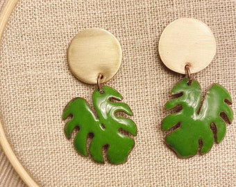 May include: A pair of gold and green earrings featuring a round gold disc and a green monstera leaf.
