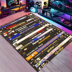 May include: A black and white rug featuring a design of VHS tapes with titles of popular horror films, including "Halloween", "Scream", "Beetlejuice", and "Friday the 13th".