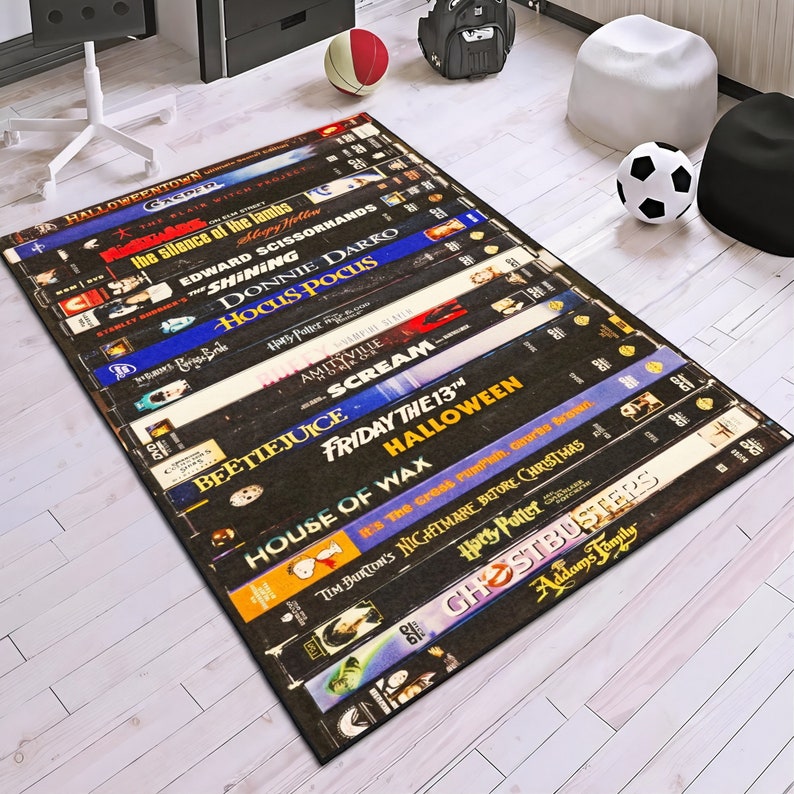 May include: A black and white rug featuring a design of stacked VHS tapes with titles of popular horror and fantasy films, including "Halloween", "Scream", "Beetlejuice", "Hocus Pocus", "The Shining", and "Nightmare Before Christmas".