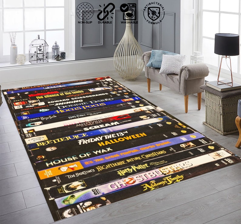 May include: A black and white rug featuring a design of stacked VHS tapes with titles of popular horror and fantasy films, including "Halloween", "Beetlejuice", "Scream", "Ghostbusters", and "The Nightmare Before Christmas".