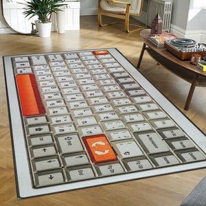 Puede incluir: Una alfombra rectangular con un diseño de teclado. La alfombra presenta un diseño de teclado gris y blanco con teclas de acento naranja. Las teclas están etiquetadas con letras, números y símbolos. La alfombra está bordeada por un marco gris fino.
