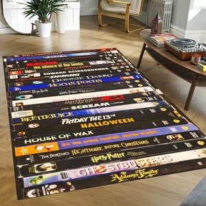 May include: A black and white rug featuring a design of classic Halloween movie VHS tapes. The tapes include titles such as "Halloween", "Beetlejuice", "Hocus Pocus", "Scream", "Friday the 13th", and "Nightmare Before Christmas".
