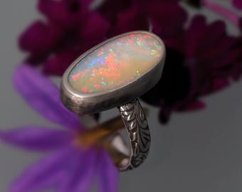 May include: Oval opal ring with a silver band. The opal displays a vibrant play of color, including red, orange, and blue hues. The ring's band features an intricate, engraved floral design. The background includes purple flowers.