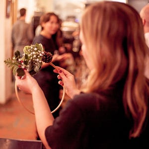 May include: A woman is making a wreath with greenery, white berries, and a pine cone. She is holding the wreath in a wooden hoop.