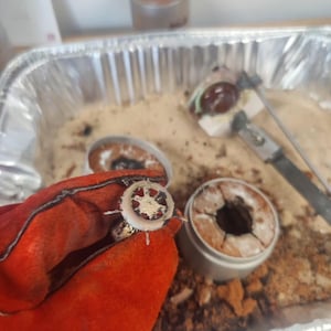 May include: A close-up of a hand wearing a red glove holding a small, circular metal object with a star-like design. Other similar objects and tools are in a metal tray filled with sand.