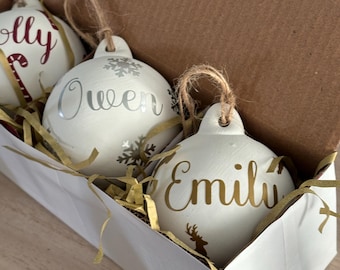 May include: A set of three white Christmas ornaments with names in gold and silver script, displayed in a white box with gold filler. Each ornament has a twine loop for hanging. One ornament has a snowflake design.