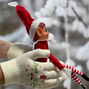 May include: A white pair of elf gloves with green text that reads "J-R Elf Gloves" and stars. The gloves are holding a red and white elf doll with a red hat.