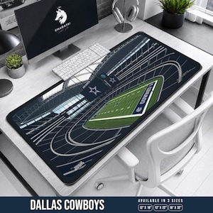 May include: A rectangular desk mat featuring an aerial graphic illustration of the Dallas Cowboys stadium with a green football field, dark blue stadium seating, and white structural roof beams. The text Dallas Cowboys is printed on the mat.