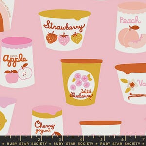 May include: A pink fabric pattern featuring stylized illustrations of yogurt containers with various fruit flavors. The containers are labeled with text such as "Strawberry," "Apple," and "Wild Blueberry." The design includes images of the fruits.