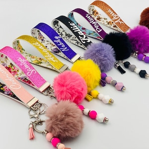 May include: A set of colorful keychains with personalized names, each featuring a fluffy pom-pom and a variety of beads. The keychains are made with glitter fabric and floral patterns.