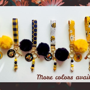 May include: A collection of bee-themed keychains with various strap designs, including yellow gingham, honeycomb, and floral patterns. Each keychain features a fluffy yellow pom-pom, a bee charm, and a round pendant with a bee illustration. The text "More colors available" is visible.