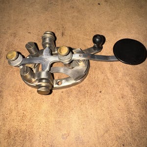 May include: A vintage telegraph key featuring a metallic base with multiple brass adjustment screws and a central cross-shaped component. The device includes a long, slender metal arm extending to the right, ending in a circular black knob.