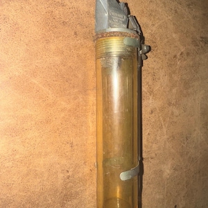 May include: A vintage, cylindrical glass oiler with a metal top and base. The clear glass tube has a slight amber tint. The metal components show signs of age and wear. The oiler has a hinged top and a small metal clip.