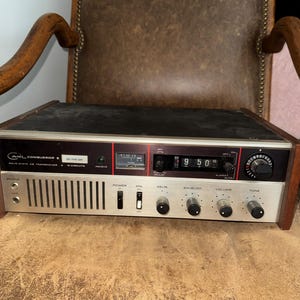 May include: A vintage Courier Conqueror II solid state CB transceiver featuring a silver front panel with black knobs, a digital clock display, a signal meter, and various switches labeled Power, ANL, Delta, Squelch, Volume, and Tone.