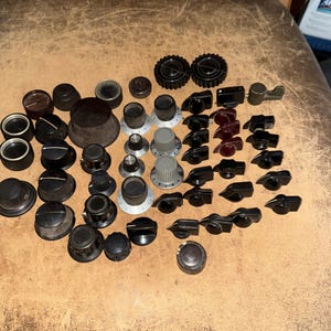 May include: A collection of various vintage electronic control knobs in black, grey, and dark red. The assortment includes round, fluted, and pointer-style knobs, some featuring numerical markings, silver-toned accents, and textured grip surfaces.