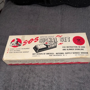 May include: Vintage S.O.S. Signal Set box, featuring the text "Official Scout Signal Set." The box includes a sturdy plastic instrument, batteries, and a code instruction booklet. The box has a red and black design with the Boy Scouts of America logo.