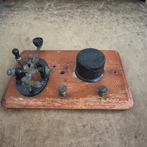May include: A vintage telegraph key mounted on a rectangular wooden base. The device features a black metal lever mechanism with adjustable screws and a cylindrical black component. Two small metal binding posts are positioned on the wood surface.