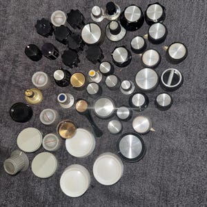 May include: A collection of various electronic control knobs in black, silver, white, and gold tones. The assortment includes circular knobs with ribbed edges, smooth flat tops, and some featuring indicator lines or numerical markings for adjustment.