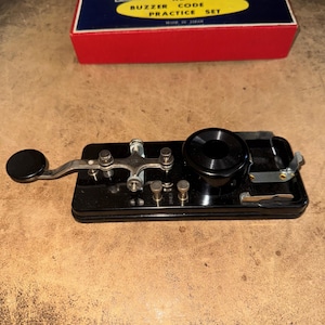 May include: A black Morse code practice key set with a black knob and a black circular sounder. The set includes a red and blue box with the text "KB-1.5 BUZZER CODE PRACTICE SET".