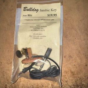 May include: A Bulldog Iambic Key model BD1 in a clear plastic bag with a paper insert. The insert displays the text Bulldog Iambic Key, Model BD1, and contact information. The key features a wooden base, metal components, and a black cable.