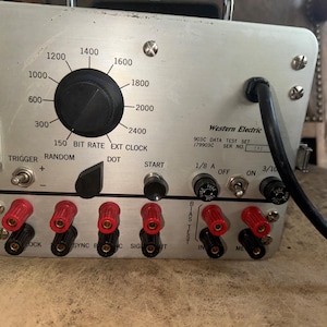 May include: A silver Western Electric 903C Data Test Set with a black knob, red and black banana plugs, and a black power cord. The device has a digital display with the numbers 1200, 1400, 1600, 1800, 2000, 2400, 150, and 300. The device also has a series of buttons labeled "Trigger", "Random", "Dot", "Start", "1/8 A", "Off", "On", "3/10", "Lock", "Sync", "Sig", "Out", "In", and "M".