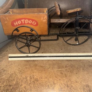 May include: A decorative wooden hotdog cart with black metal wheels and frame. The cart features the word "HOTDOG" in red on the side. The cart has a wooden handle and seat. A ruler is placed in front of the cart.