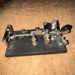 May include: A vintage telegraph key featuring a black rectangular base with a textured finish. The device includes polished chrome metal components, adjustable screws, a horizontal lever arm, and a black circular knob at the end for operation.