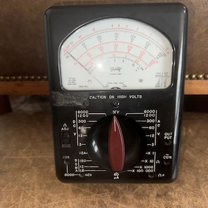 May include: A black analog multimeter with a white face and a red needle. The dial has markings for volts, amps, ohms, and decibels. The device has a red knob and several switches labeled "ADJ", "OUT PUT", and "COM".