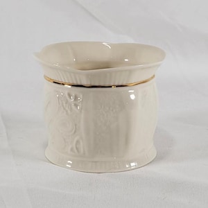 May include: Cream-colored ceramic vase with a scalloped rim and gold trim. The vase features embossed floral and scrollwork designs. The base is slightly flared, and the overall shape is cylindrical.