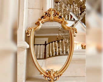 May include: A gold ornate mirror with a shield-shaped frame. The mirror is reflecting a white staircase with a gold railing.