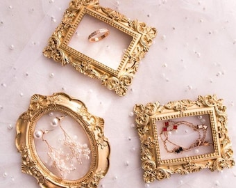 May include: Three ornate gold-colored frames with glass inserts for displaying jewelry. The frames are decorated with intricate details and have a vintage aesthetic.