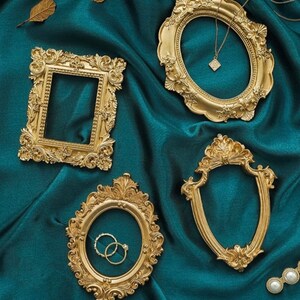 May include: Four ornate gold-colored frames with intricate details, set against a teal fabric background. The frames are different shapes and sizes, and one frame has a necklace inside.