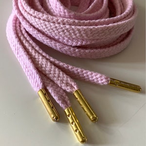 May include: Close-up of a bundle of light pink shoelaces with gold-colored aglets. The laces are made of a woven material and are neatly coiled. The aglets have small holes. The laces are on a white surface.