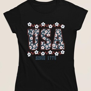 May include: Black t-shirt with the word "USA" in a floral pattern, outlined in red and white. The text "SINCE 1776" is below the word "USA". The shirt has short sleeves and a round neckline.