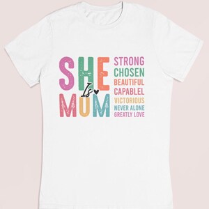 May include: White t-shirt with a colorful graphic that says "She is Mum" with the words "Strong, Chosen, Beautiful, Capable, Victorious, Never Alone, Greatly Love" stacked vertically.