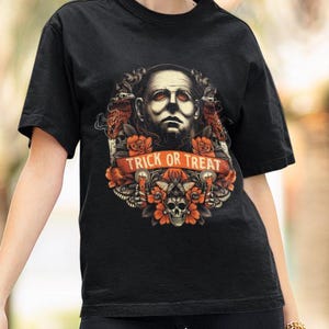 May include: Black t-shirt featuring a Halloween-themed graphic. The design includes a central image of a person's face, surrounded by orange flowers, skulls, and the text "TRICK OR TREAT" in a banner. The shirt has short sleeves.