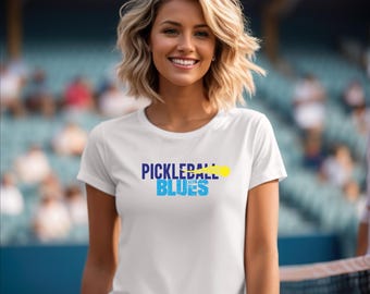Może przedstawiać: Biała koszulka z niebiesko-żółtym nadrukiem z napisem "Pickleball Blues".